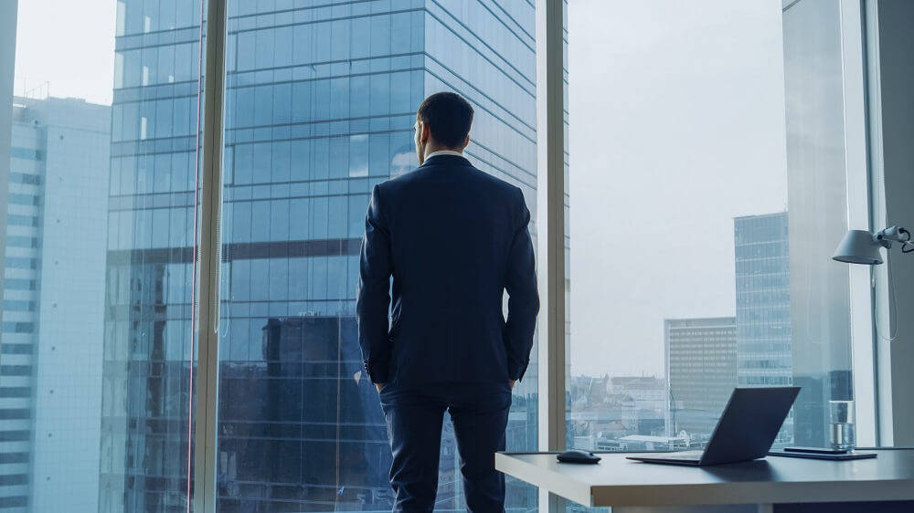 man in suit standing next to large window from an office building looking contemplative