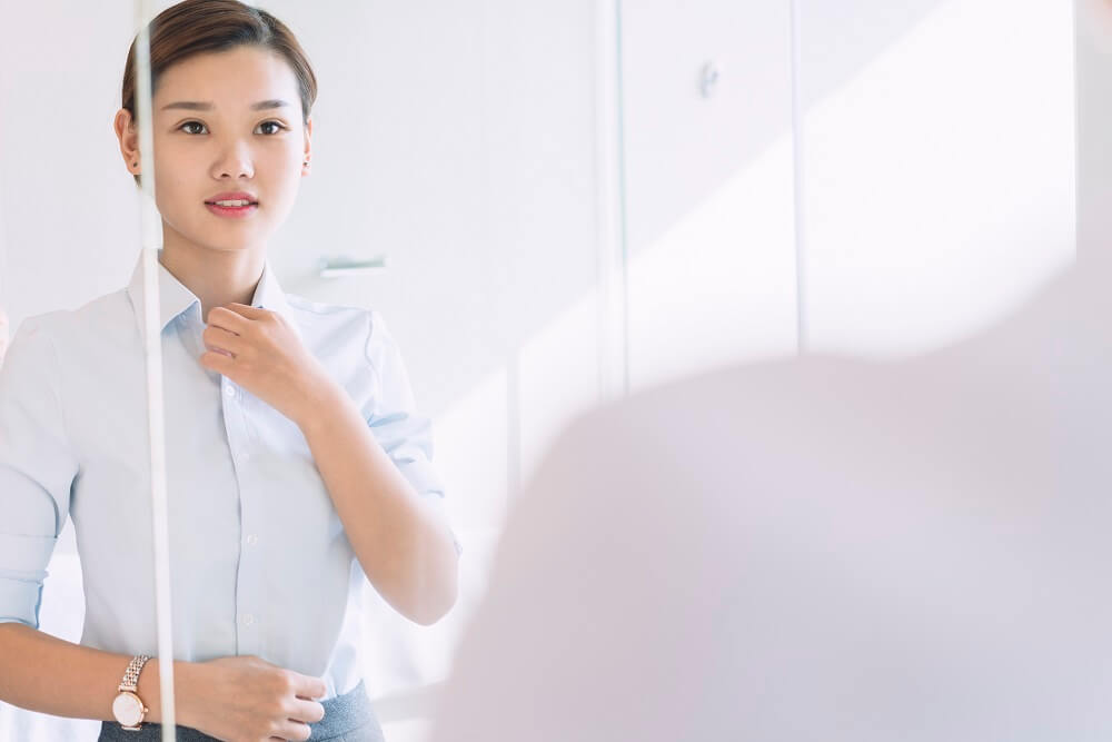 east asian woman with brown hair in a bun looking into a mirror and buttoning her shirt preparing for an interview