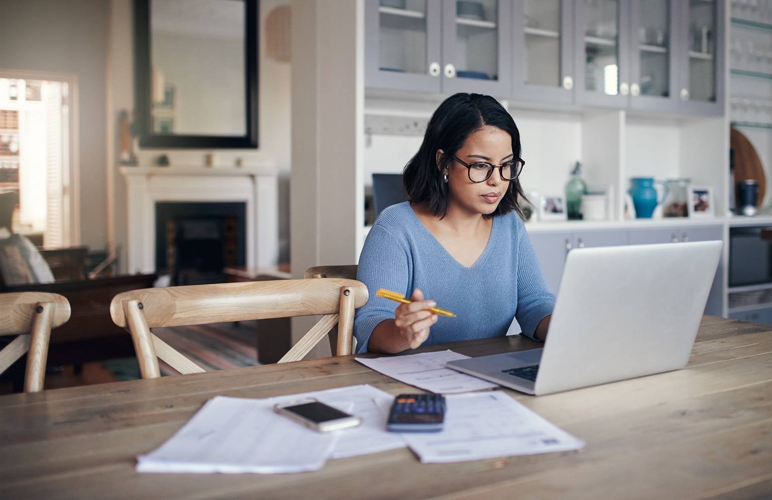 asian woman in glasses working at her kitchen table with her laptop and papers 